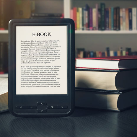 E-book reader displaying text on a table in front of stacked physical books, with a bookshelf in the background