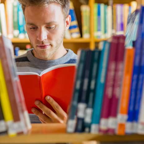 Student reading an orange book in a library, surrounded by shelves filled with colorful books