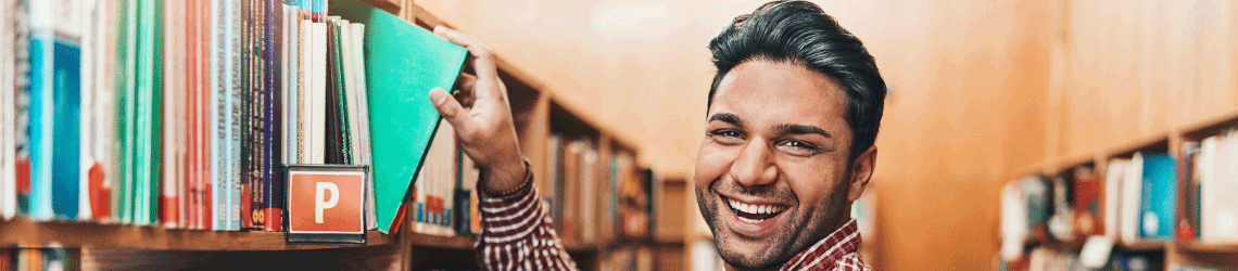 a student smiling while pulling out a book from a library shelf