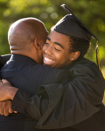 A college graduate smiles as he hugs his parent while wearing his college regalia