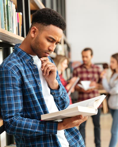 A student leans against a bookcase and reads a book