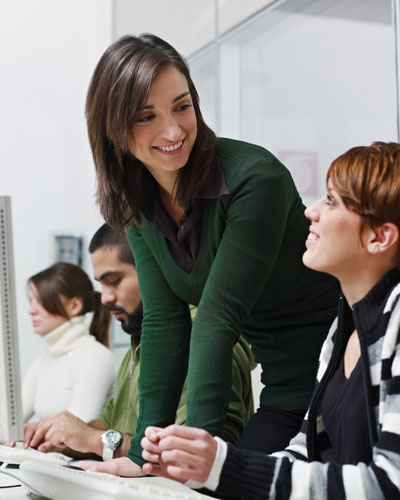 Two women talk near a computer. One is seated and the other is standing.