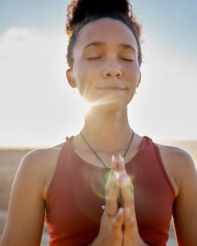 A woman meditates with her hands clasped and the sun setting behind her