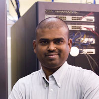 A person in a light gray shirt stands confidently in a server room, with racks of computing equipment behind them