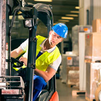 A worker wearing a blue hard hat and yellow vest operates a forklift in a warehouse.