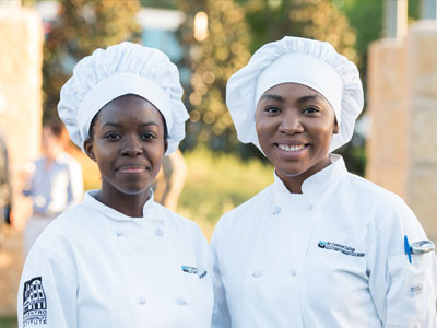 Two chefs in white uniforms and hats stand smiling outdoors
