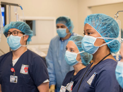 Healthcare professionals in blue scrubs, masks, and hairnets attentively listen in a hospital setting