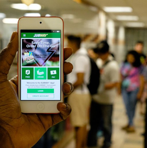 A person holds up a Subway mobile app while students stand in the background