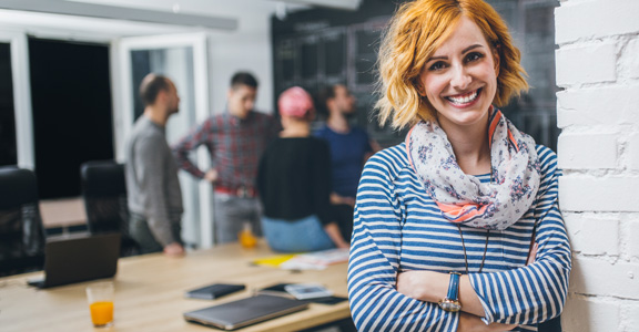 An office worker stands in the foreground with her arms crossed while her co-workers discuss something in the background.