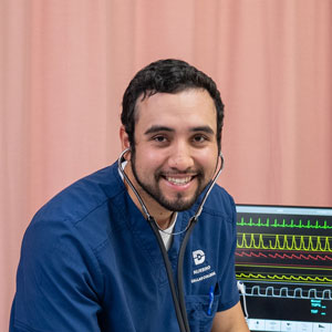 A nursing student sits next to a bed with a monitor running in the background