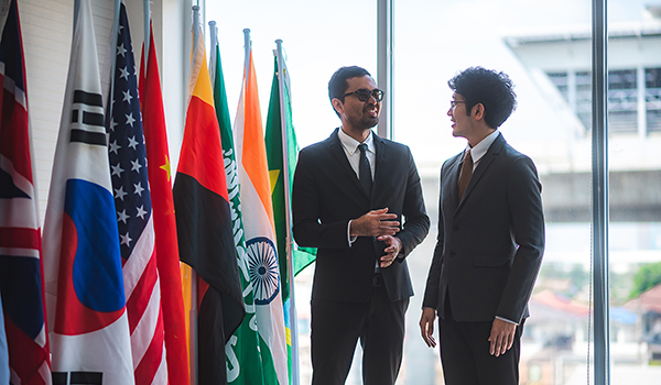 Two men dressed in suits talk to one another in front of a row of international flags
