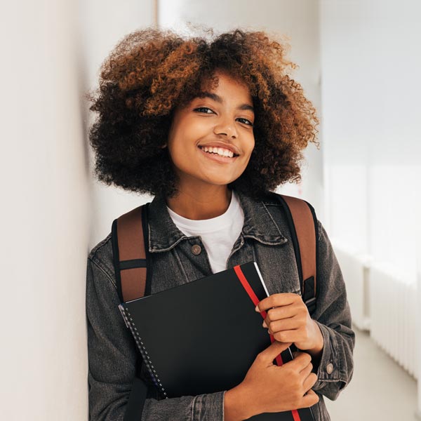A student holds a notebook in a hallway while smiling towards the camera