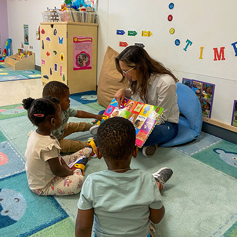 A teacher sits on the floor of a classroom reading to three young children