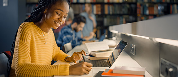 female student sitting at desk using laptop and writing notes