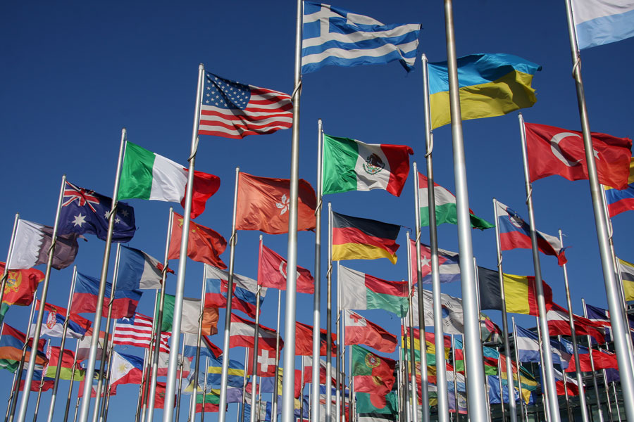Multiple national flags, including the U.S., Greece, Ukraine, Turkey, Germany, and Mexico, waving on flagpoles against a clear blue sky.