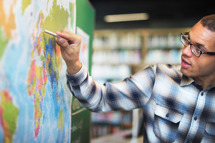 Person in plaid shirt pointing at a colorful world map with a pen; blurred bookshelves in the background suggest a library setting.