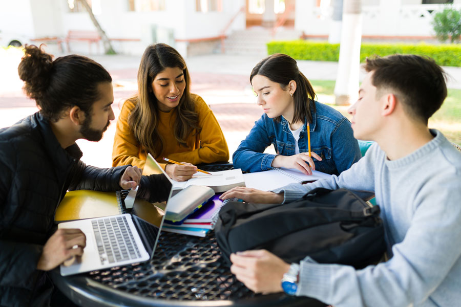 A group of four students sitting around a round outdoor table, engaged in a study session. They have books, notebooks, and a laptop on the table. The background shows a campus-like setting with greenery and buildings.
