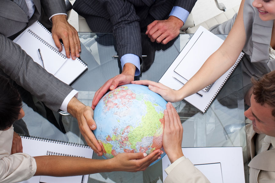Group of professionals in business attire seated around a glass table, reaching toward a large globe at the center, symbolizing global collaboration.