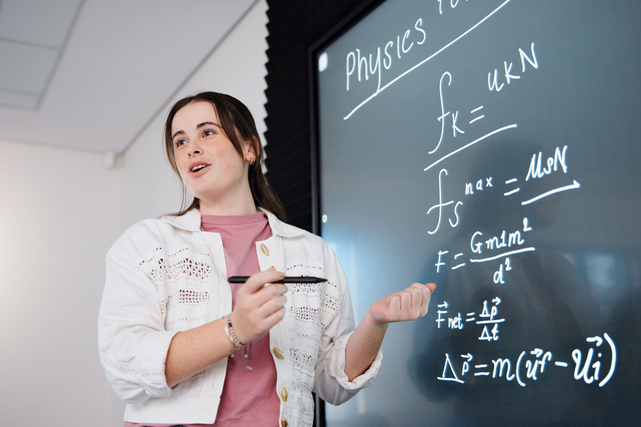 Student standing in front of a blackboard with physics equations, holding a marker and gesturing.