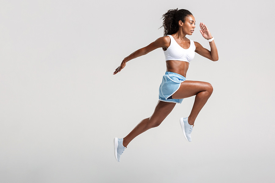 Person in athletic wear mid-jump against a plain background, wearing a white sports bra and light blue shorts.