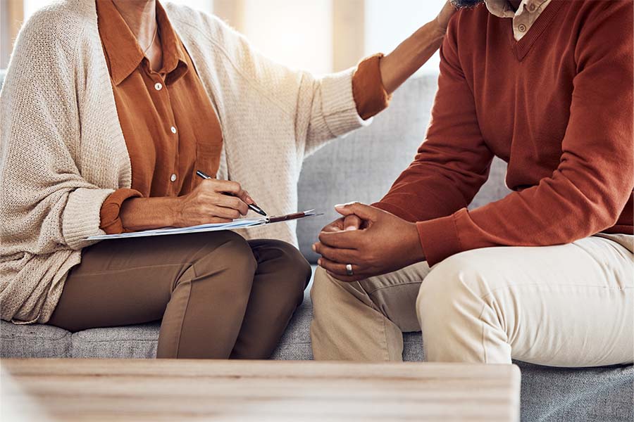 Person in beige cardigan holds a clipboard and pen; another in orange sweater sits with hands clasped.