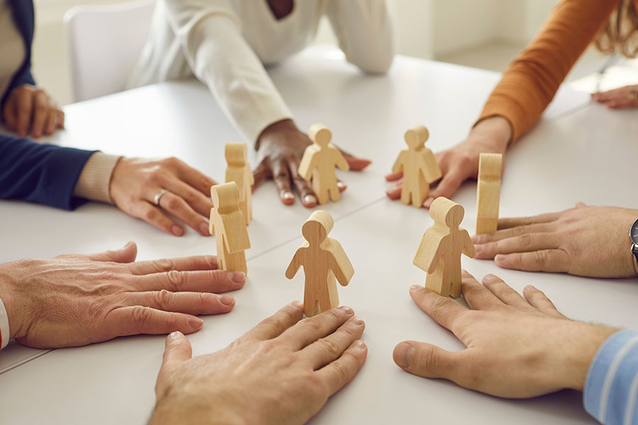 Group of people sit around a white table with hands placed near wooden figurines arranged in a circle, symbolizing teamwork.