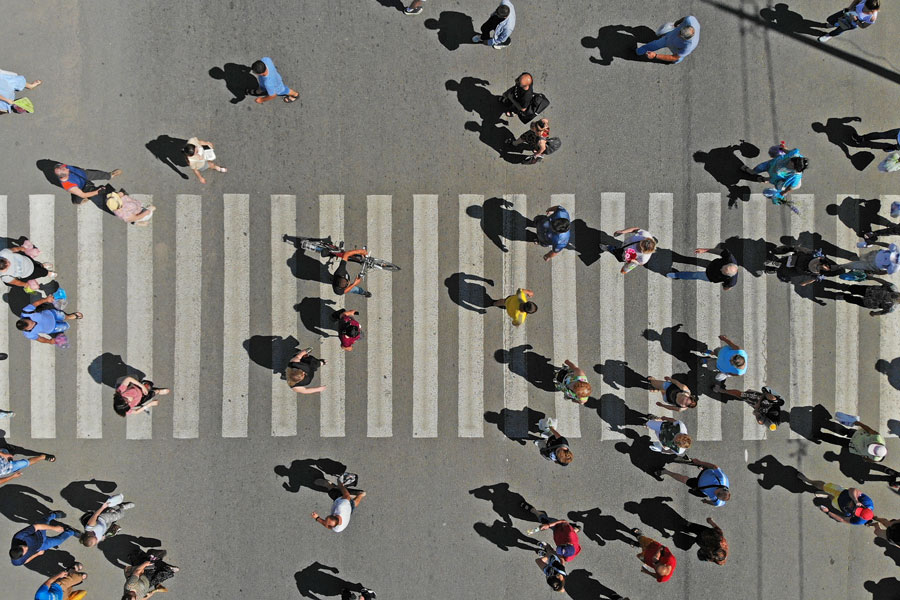 Aerial view of a crosswalk with people walking in various directions; white stripes on gray road with visible shadows from sunlight.